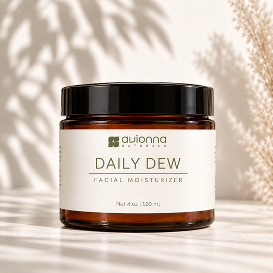 A jar of Avionna Naturals Daily Dew Facial Moisturizer, featuring an amber glass jar with a black lid and a white label, set against a warm beige background with soft leaf shadows.