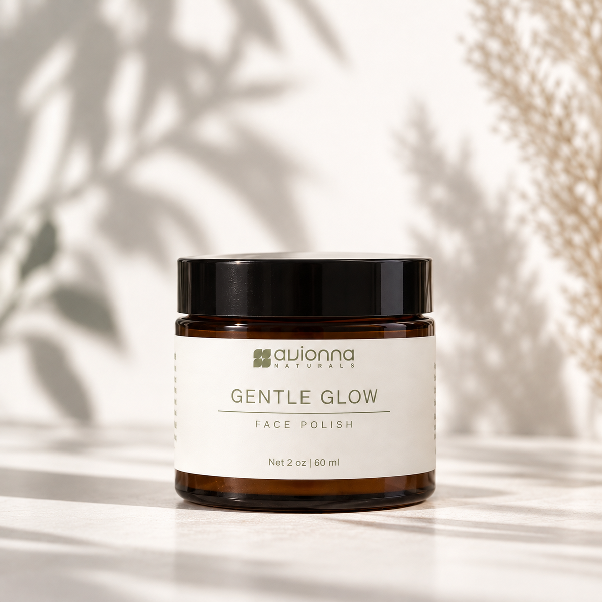 A jar of Avionna Naturals Gentle Glow Face Polish sits on a light surface with soft leaf shadows and dried grass in the background. The product is in an amber glass jar with a black lid and a white label.
