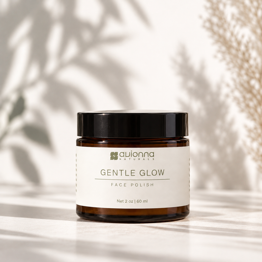 A jar of Avionna Naturals Gentle Glow Face Polish sits on a light surface with soft leaf shadows and dried grass in the background. The product is in an amber glass jar with a black lid and a white label.